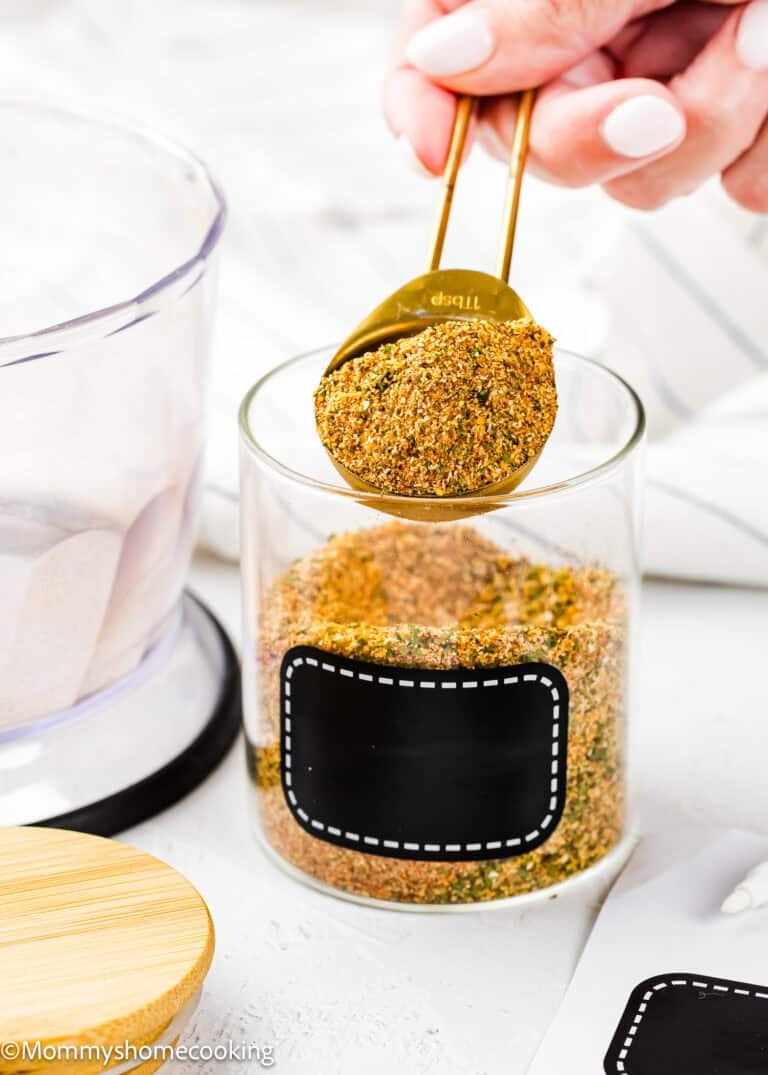 A hand holds a tablespoon of Easy Homemade Everyday Seasoning (All-Purpose & Allergy-Safe) over a clear glass jar filled with the same blend; the jar has a blank black label.