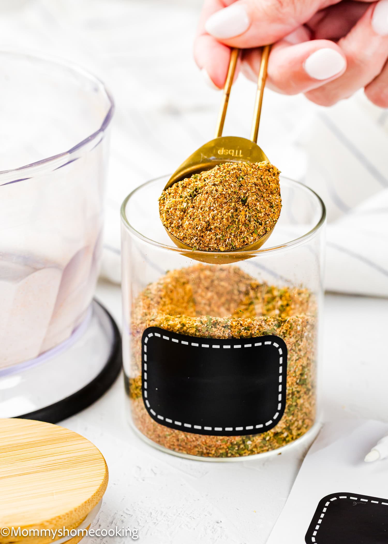 A hand holds a tablespoon of Easy Homemade Everyday Seasoning (All-Purpose & Allergy-Safe) over a clear glass jar filled with the same blend; the jar has a blank black label.