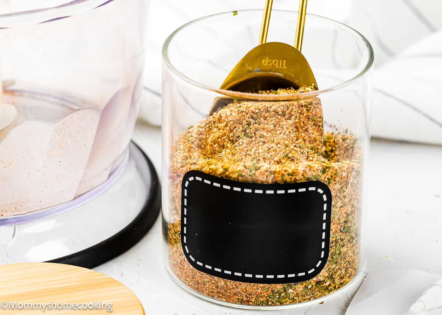 A glass jar filled with Easy Homemade Everyday Seasoning (All-Purpose & Allergy-Safe) and a gold measuring spoon, featuring a blank black label on the jar.