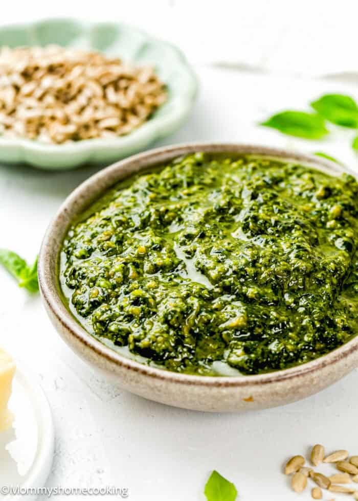 A bowl of Easy Nut-Free Pesto sauce on a white surface, with a dish of sunflower seeds and a piece of cheese nearby.