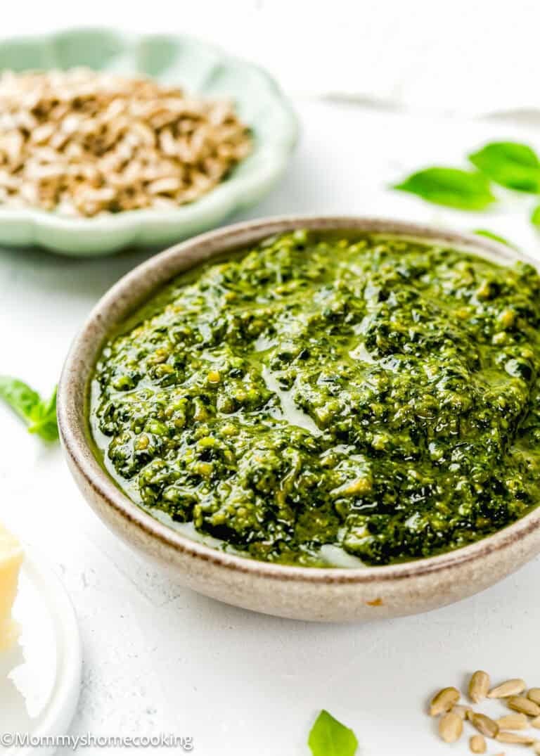 A bowl of Easy Nut-Free Pesto sauce on a white surface, with a dish of sunflower seeds and a piece of cheese nearby.