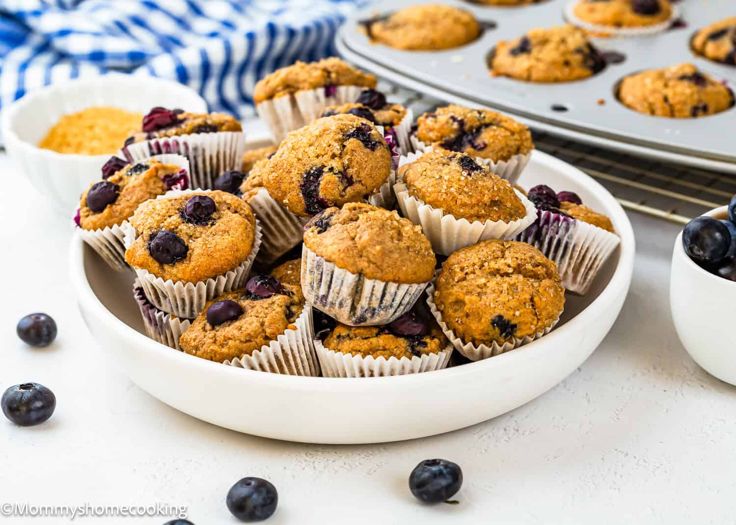 A white bowl filled with Dairy-Free Mini Blueberry Muffins (Kid-Approved + Freezer-Friendly!) in paper liners, with more Egg-Free muffins on a cooling rack and a muffin tray in the background. Fresh blueberries are scattered on the table.