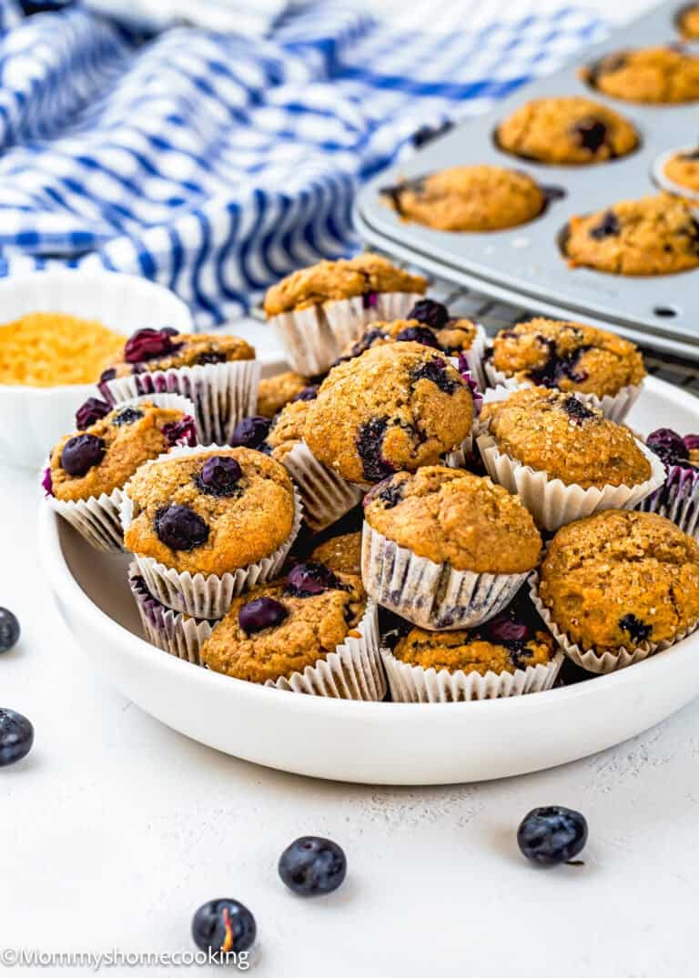 A white plate filled with Dairy-Free Mini Blueberry Muffins (Kid-Approved + Freezer-Friendly!) sits on a white surface, with more egg-free muffins in a tin and fresh blueberries scattered around.
