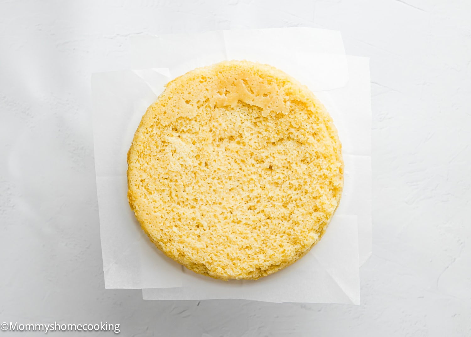 A single round Egg-Free Vanilla Cake layer sits on a piece of parchment paper on a white surface, showing off its soft, fluffy texture perfect for a birthday cake without eggs.