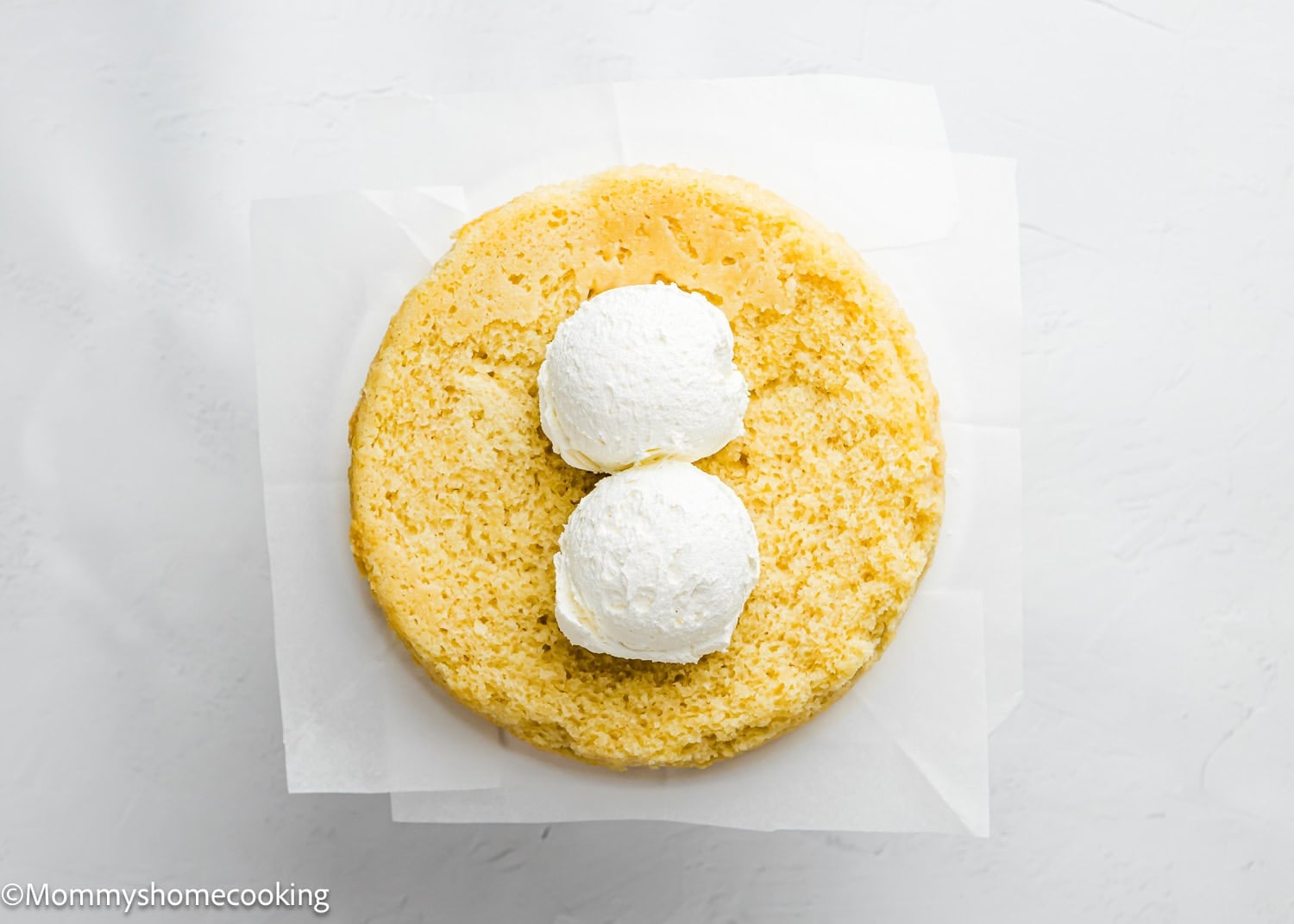 A round Egg-Free Vanilla Cake layer with two scoops of white frosting on top, placed on parchment paper over a white surface. This soft, fluffy birthday cake is made without eggs.