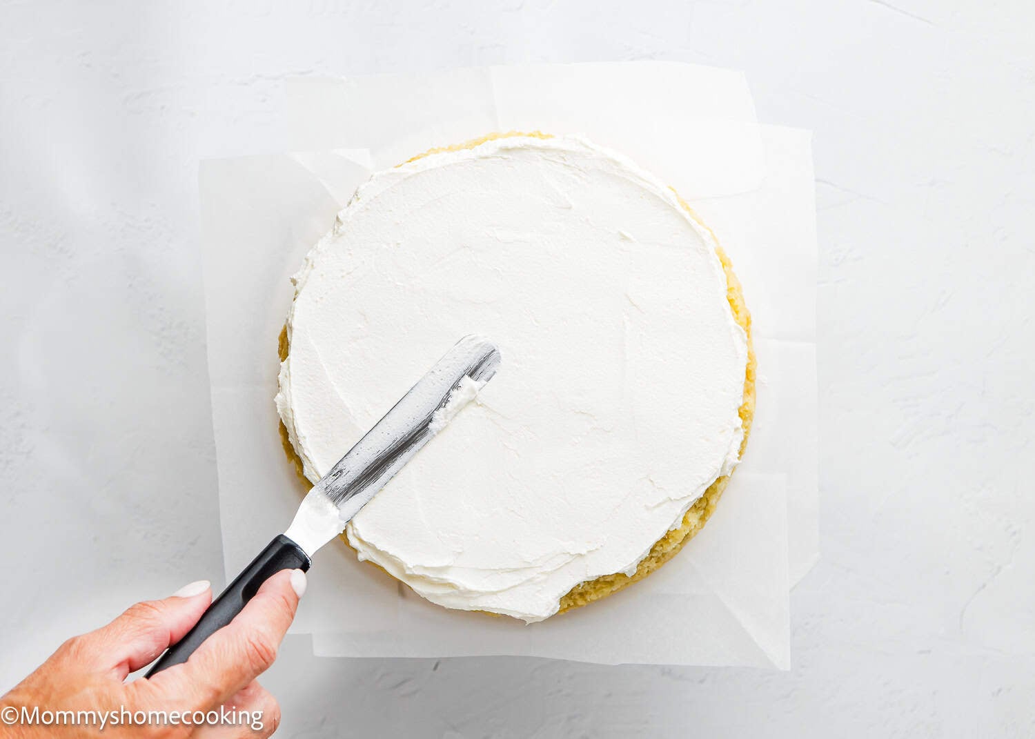 A hand spreads white frosting evenly over an Egg-Free Vanilla Cake, a soft, fluffy birthday cake without eggs, using an offset spatula on parchment paper.