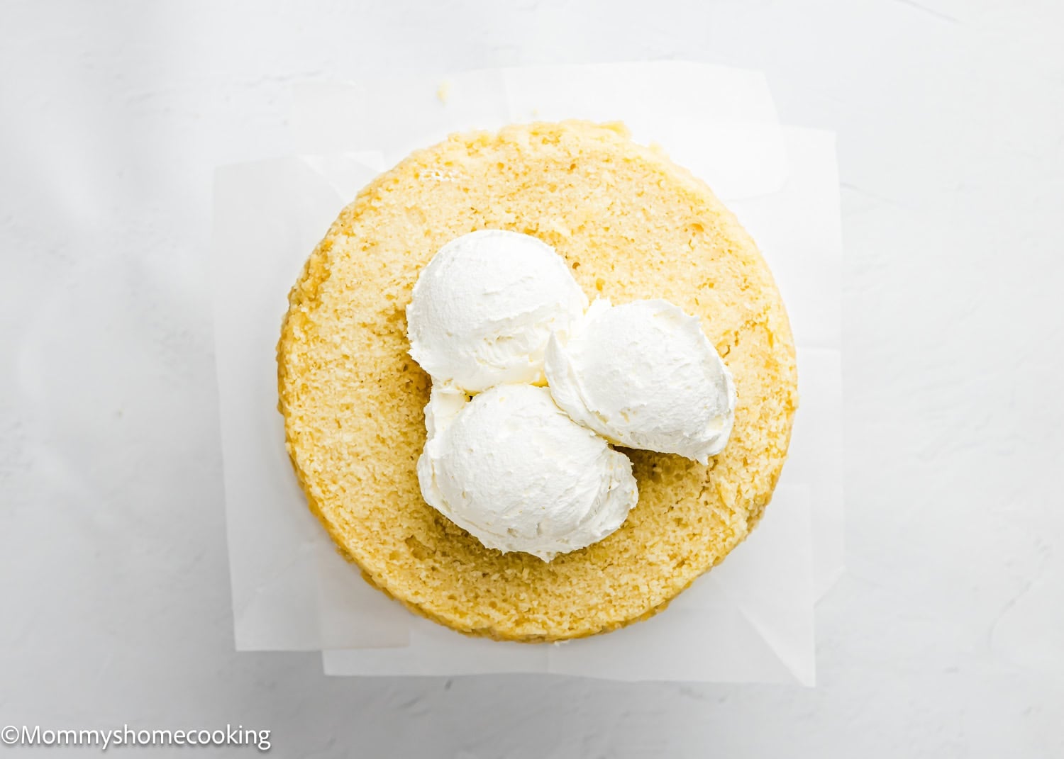 A plain round Egg-Free Vanilla Cake – soft, rests on parchment paper with three large scoops of white frosting on top, ready to be spread.