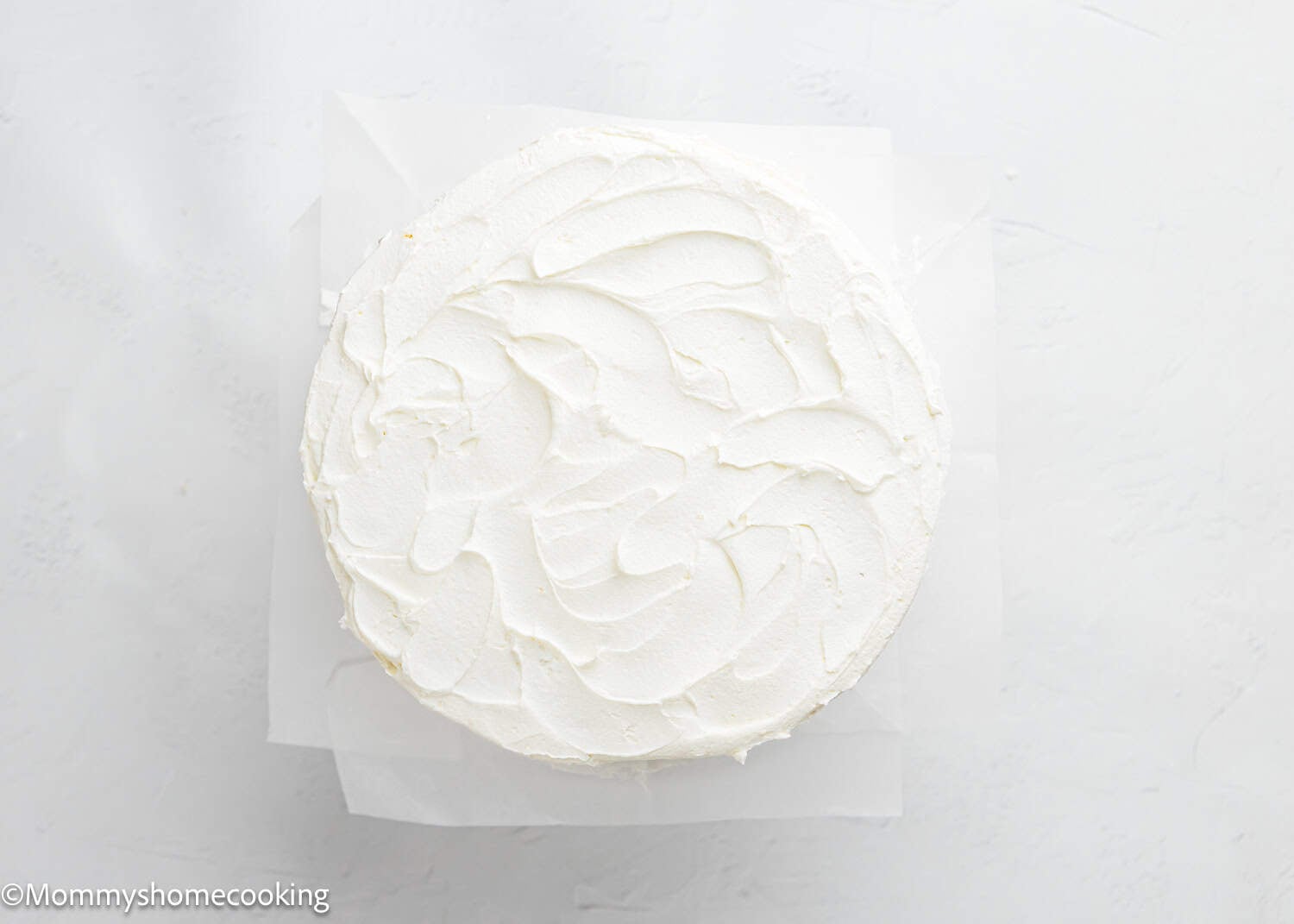 A fluffy birthday cake without eggs, covered with smooth white frosting, sits on a parchment-lined surface and is viewed from above.