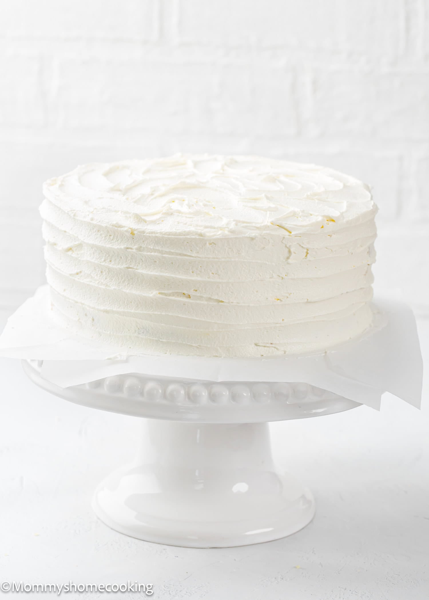 A round, white frosted Egg-Free Vanilla Cake sits on a white cake stand against a white background. This soft, fluffy birthday cake is made without eggs.