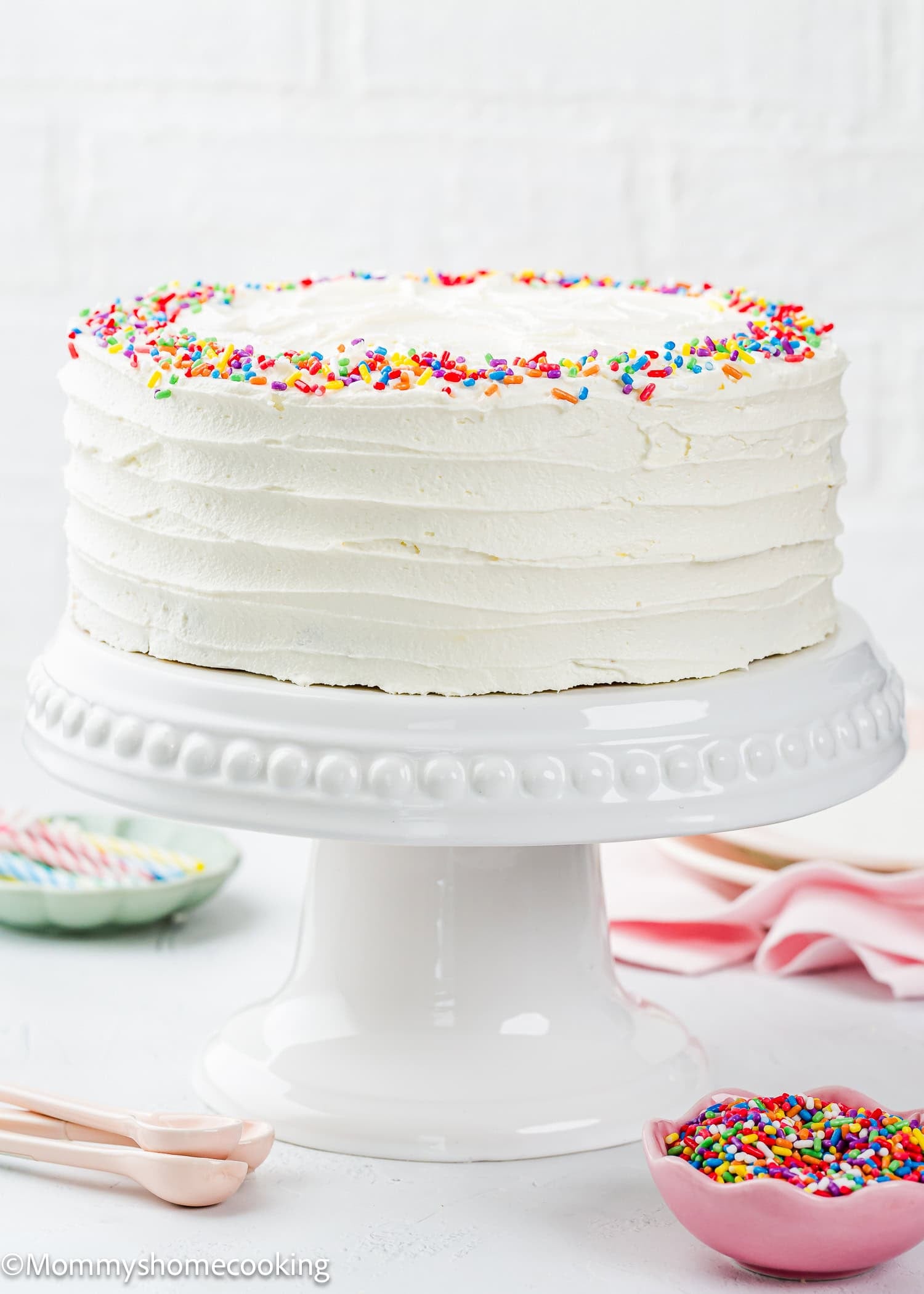 A round white frosted Egg-Free Vanilla Cake with colorful sprinkles sits on a white cake stand, surrounded by small bowls and pink utensils. This soft, fluffy birthday cake is made without eggs.