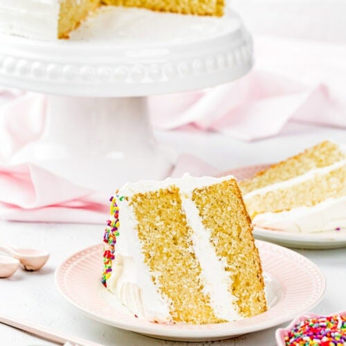 A slice of Egg-Free Vanilla Cake with white frosting and sprinkles is served on a pink plate; the rest of this soft, fluffy birthday cake without eggs sits on a stand in the background.