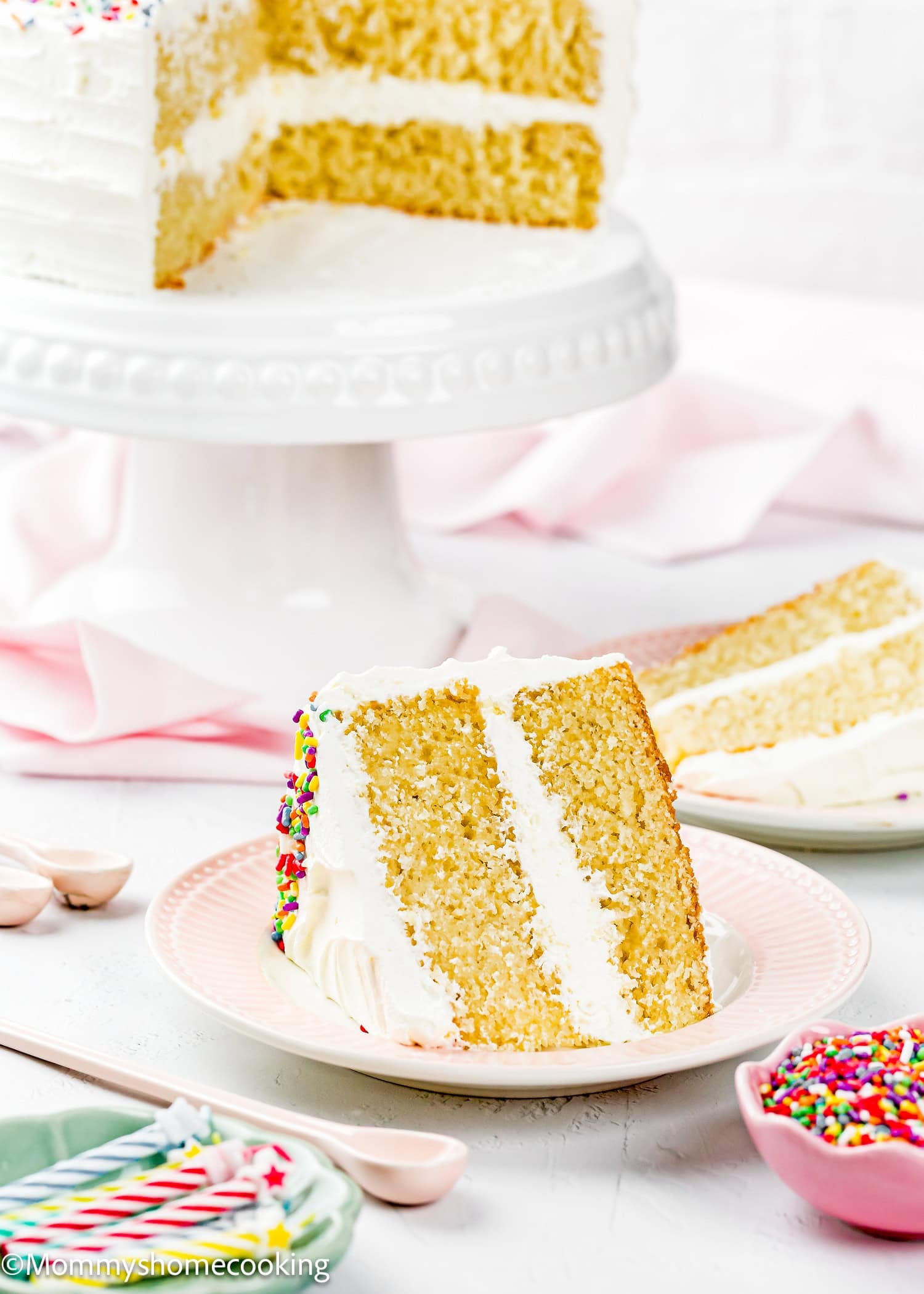 A slice of Egg-Free Vanilla Cake with white frosting and sprinkles is served on a pink plate; the rest of this soft, fluffy birthday cake without eggs sits on a stand in the background.
