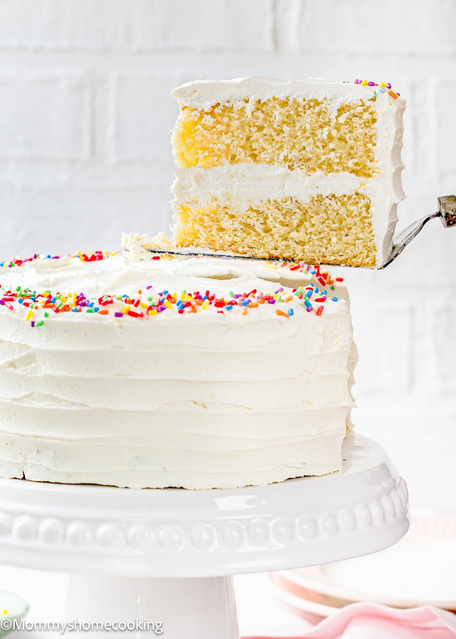 A slice of soft, fluffy Egg-Free Vanilla Cake with white frosting and rainbow sprinkles is being lifted from the whole cake on a white cake stand. This delicious birthday cake is made without eggs.