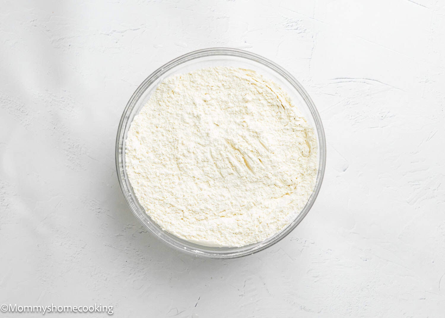 A glass bowl filled with dry ingredients over a white colored surface, perfect for baking a soft, fluffy Egg-Free Vanilla Cake without eggs.