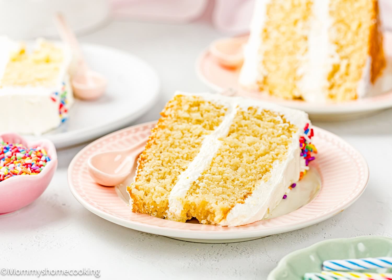 A slice of Egg-Free Vanilla Cake with white frosting and sprinkles sits on a pink plate; more cake and a bowl of sprinkles are in the background, showcasing this soft, fluffy birthday cake without eggs.