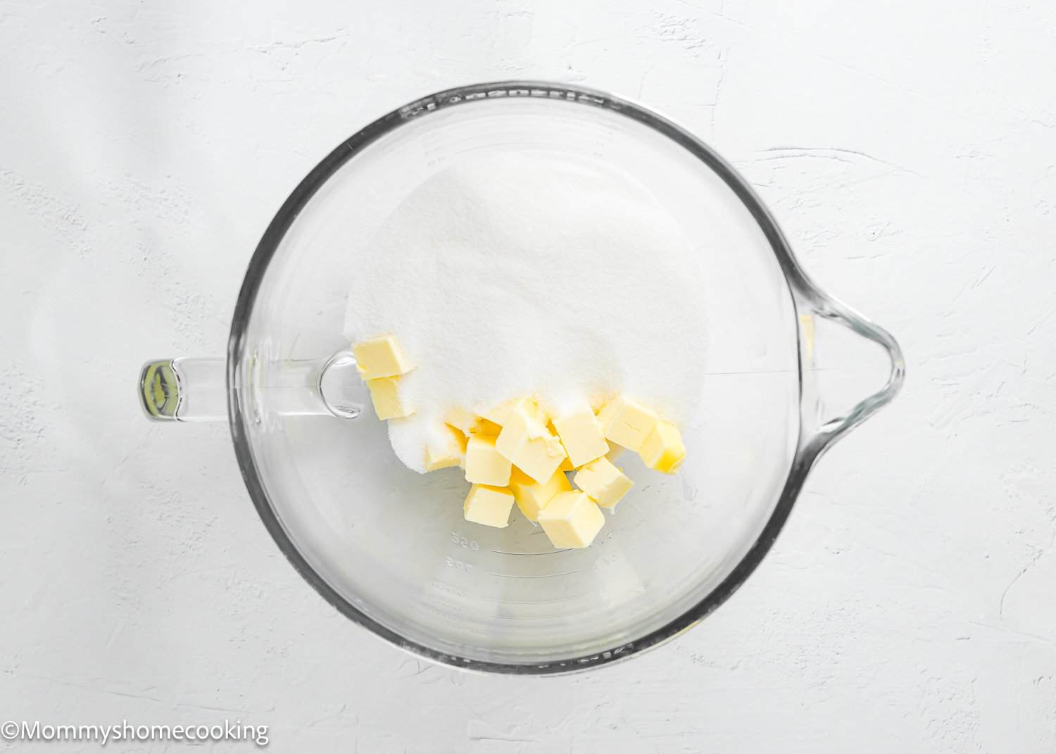 A glass mixing bowl containing white granulated sugar and cubed butter on a white surface, perfect for making a fluffy birthday cake without eggs or an Egg-Free Vanilla Cake – soft and delicious.