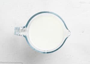A glass measuring cup filled with milk, perfect for making a fluffy birthday cake without eggs, sits on a white textured surface.