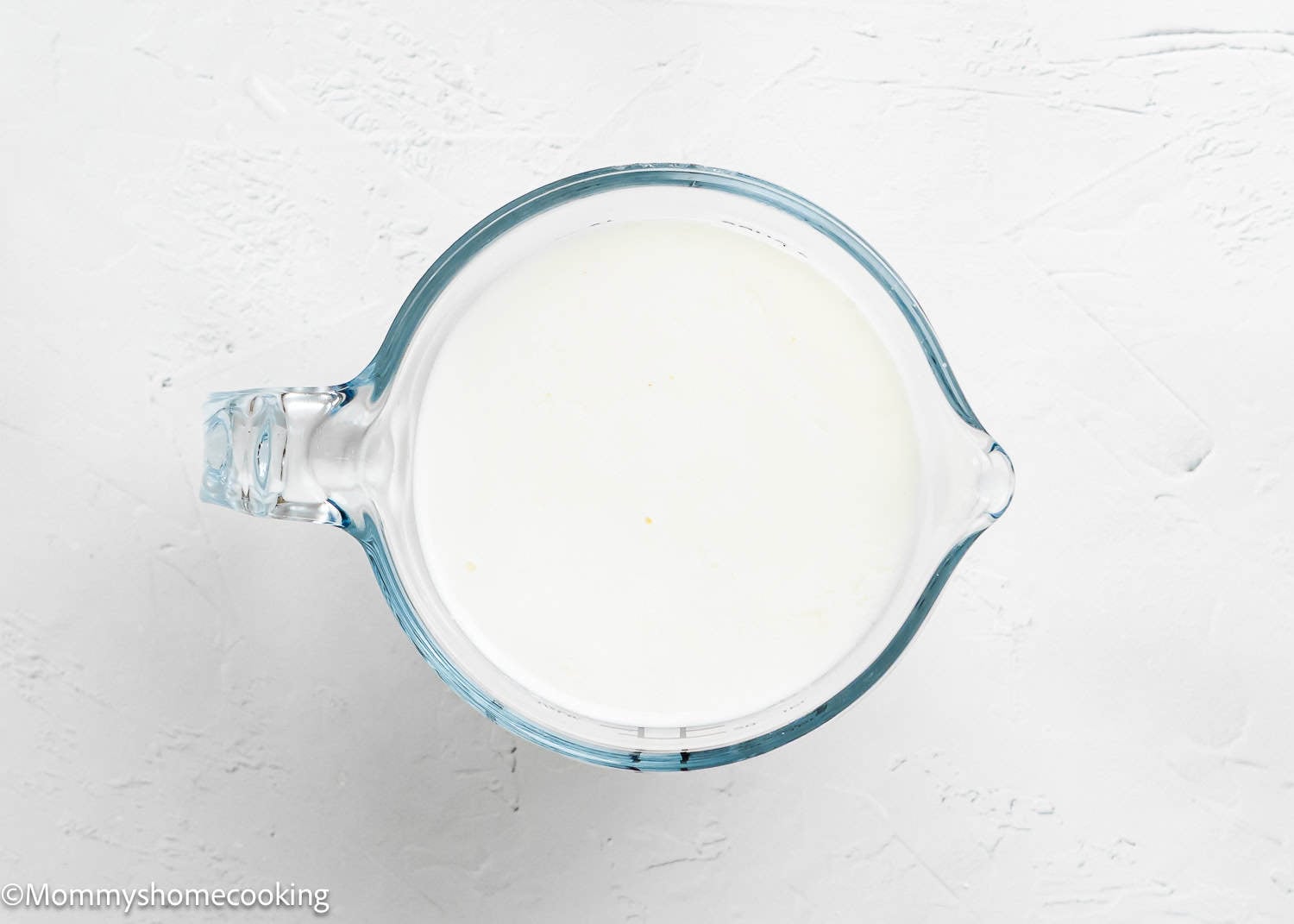 A glass measuring cup filled with milk and vinegar, perfect for making a fluffy birthday cake without eggs, sits on a white textured surface.