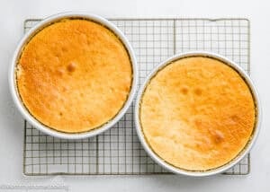 Two round, golden-brown Egg-Free Vanilla Cakes in white baking pans are cooling on a wire rack against a white background. These soft, fluffy birthday cakes are made without eggs.