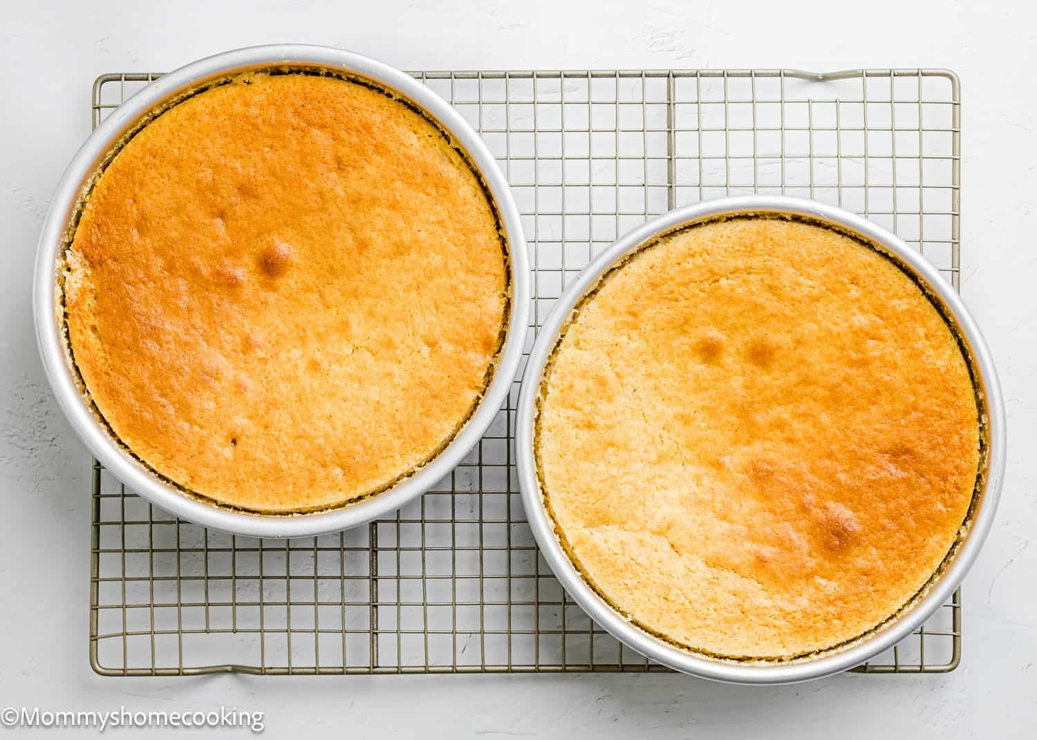 Two round, golden-brown Egg-Free Vanilla Cakes in white baking pans are cooling on a wire rack against a white background. These soft, fluffy birthday cakes are made without eggs.