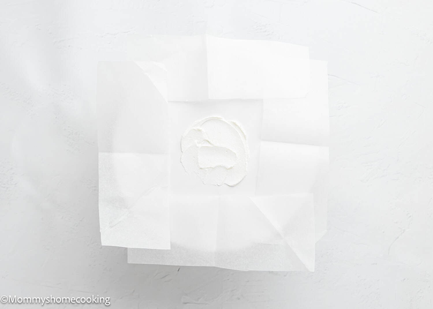 A small amount of dough for an egg-free vanilla cake sits in the center of a plate, covered by overlapping sheets of white parchment paper.