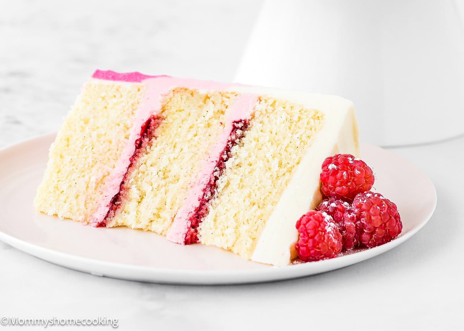 A slice of Eggless Raspberry Vanilla Layer Cake with pink and red filling, served on a white plate with fresh raspberries on the side.