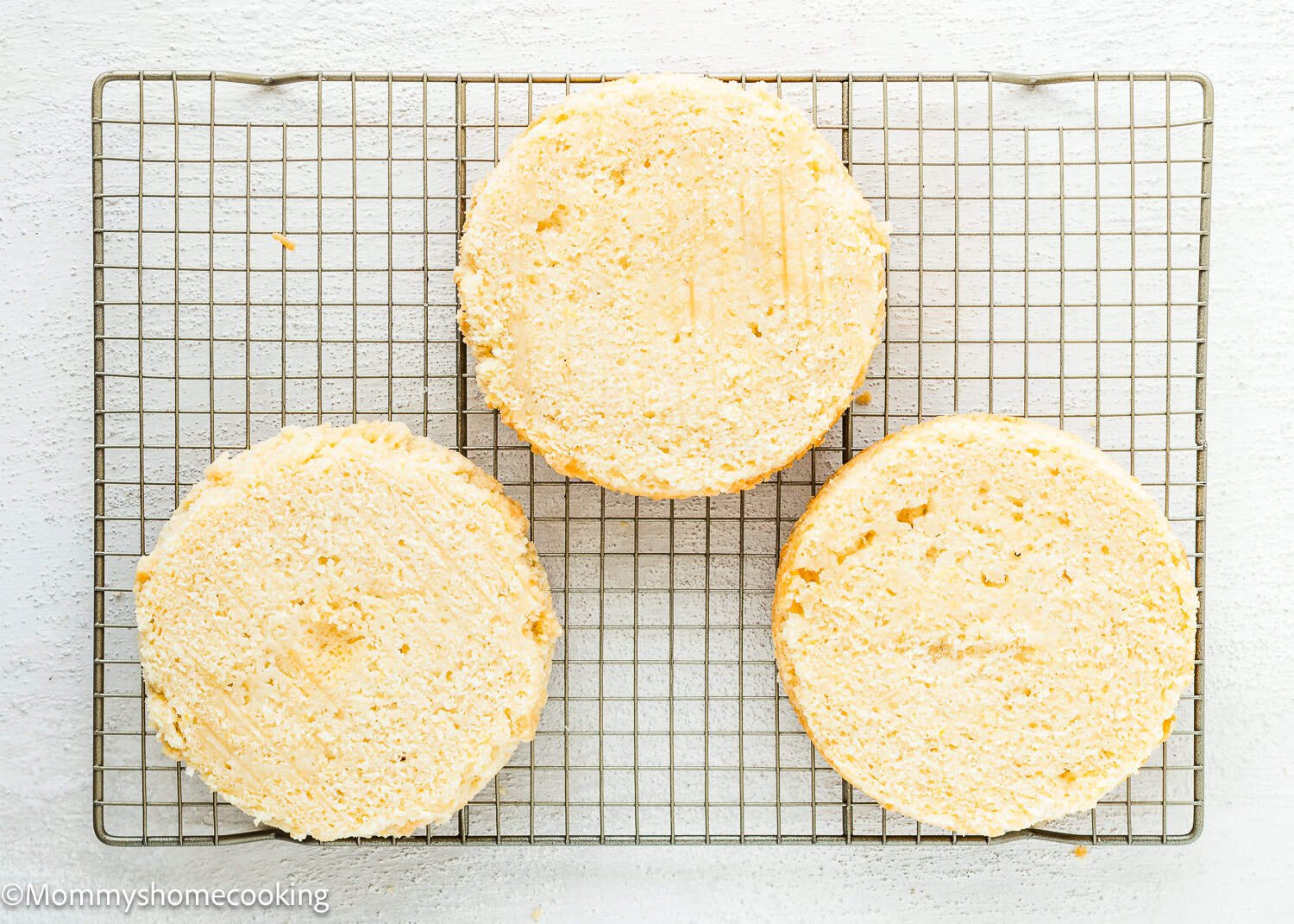 Three round layers of Eggless Raspberry Vanilla Layer Cake are cooling on a metal wire rack over a white surface.