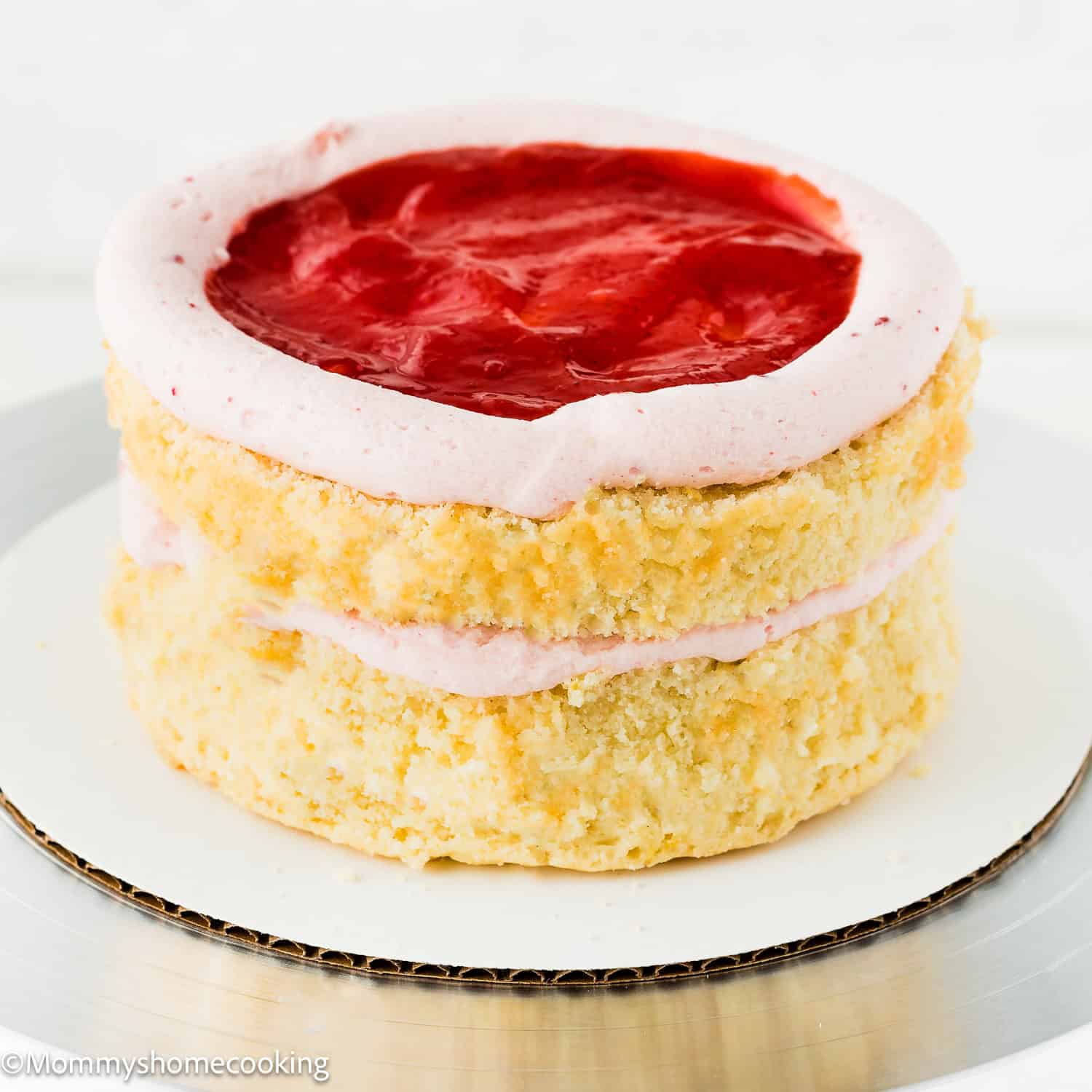 An Eggless Raspberry Vanilla Layer Cake featuring two layers of vanilla cake, pink frosting, and a layer of red strawberry topping on the upper layer, displayed on a white circular base.