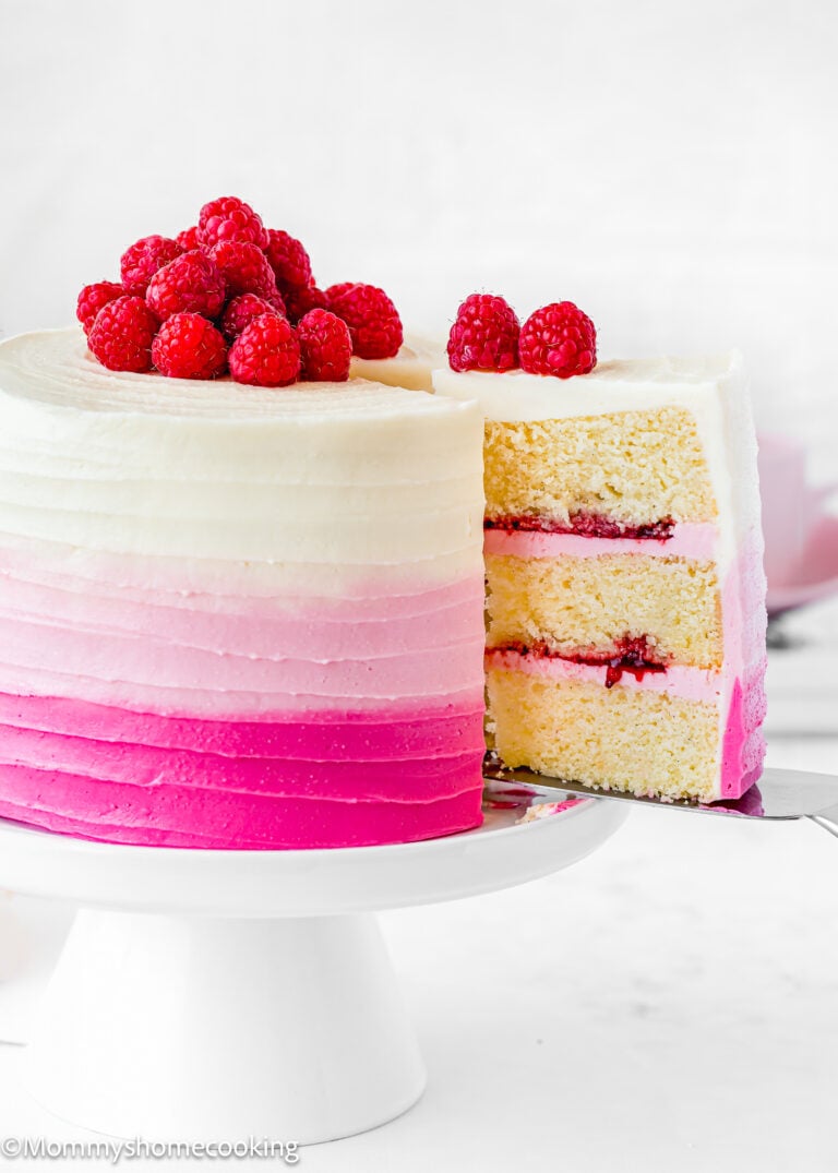 An Eggless Raspberry Vanilla Layer Cake with pink and white ombre frosting, topped with fresh raspberries, sits on a white cake stand with one slice removed.
