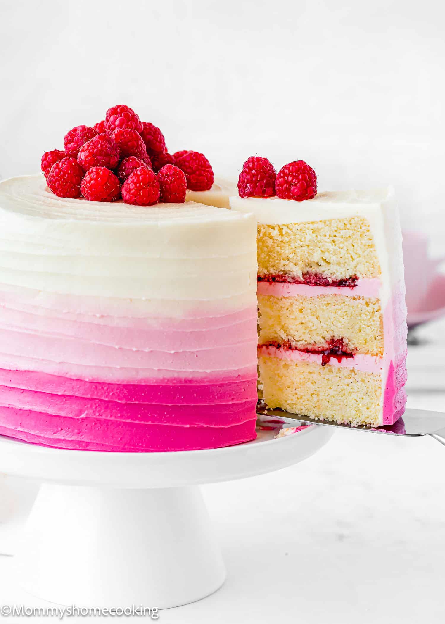 An Eggless Raspberry Vanilla Layer Cake with pink and white ombre frosting, topped with fresh raspberries, sits on a white cake stand with one slice removed.