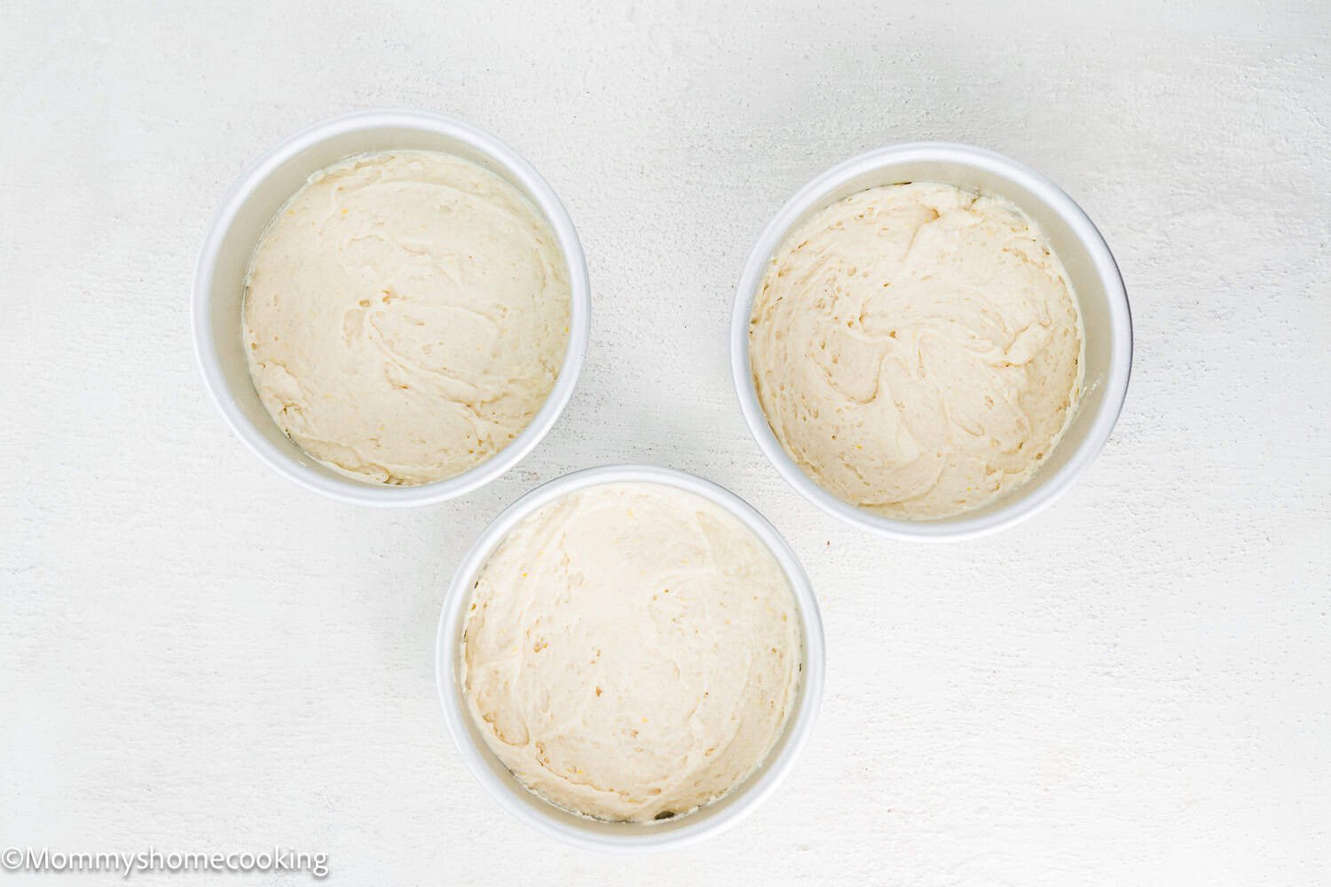 Three white cake pans filled with creamy Eggless Raspberry Vanilla Bean Layer Cake batter, arranged in a triangle on a white textured surface.