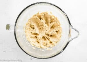 A glass mixing bowl containing creamed dough for Eggless S’mores Cookie Bars sits on a white surface.