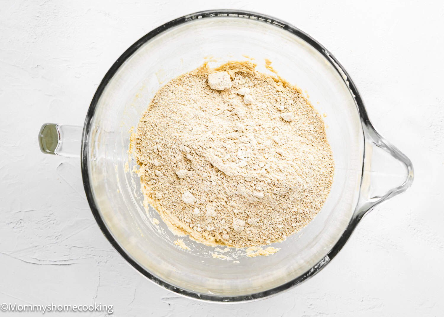 A glass mixing bowl contains a mound of dry, light brown flour mixture on a white surface, ready to create Eggless S’mores Cookie Bars.