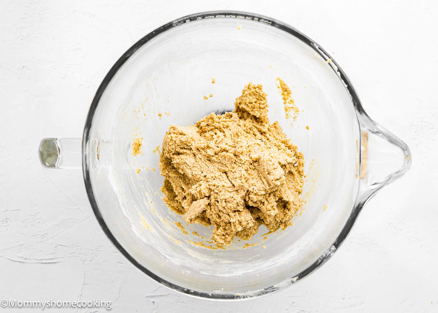 A glass mixing bowl containing a portion of Eggless S’mores Cookie Bars dough on a white surface.