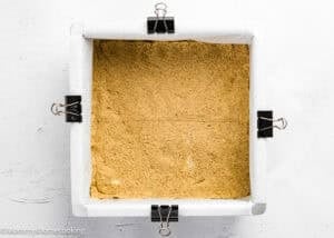 Square baking pan lined with parchment paper, secured with binder clips, containing an even layer of unbaked crust for Eggless S’mores Cookie Bars.