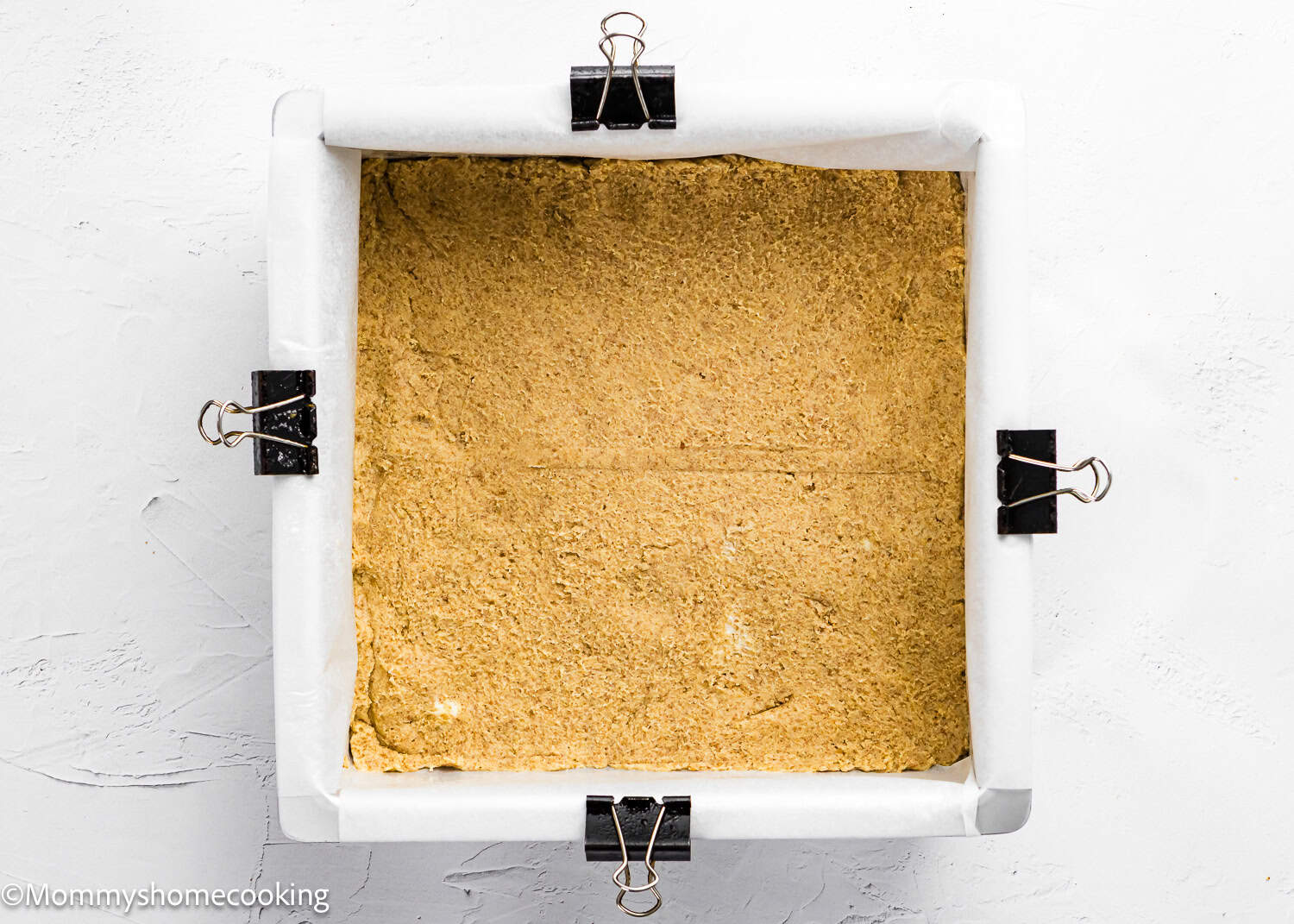 Square baking pan lined with parchment paper, secured with binder clips, containing an even layer of unbaked crust for Eggless S’mores Cookie Bars.