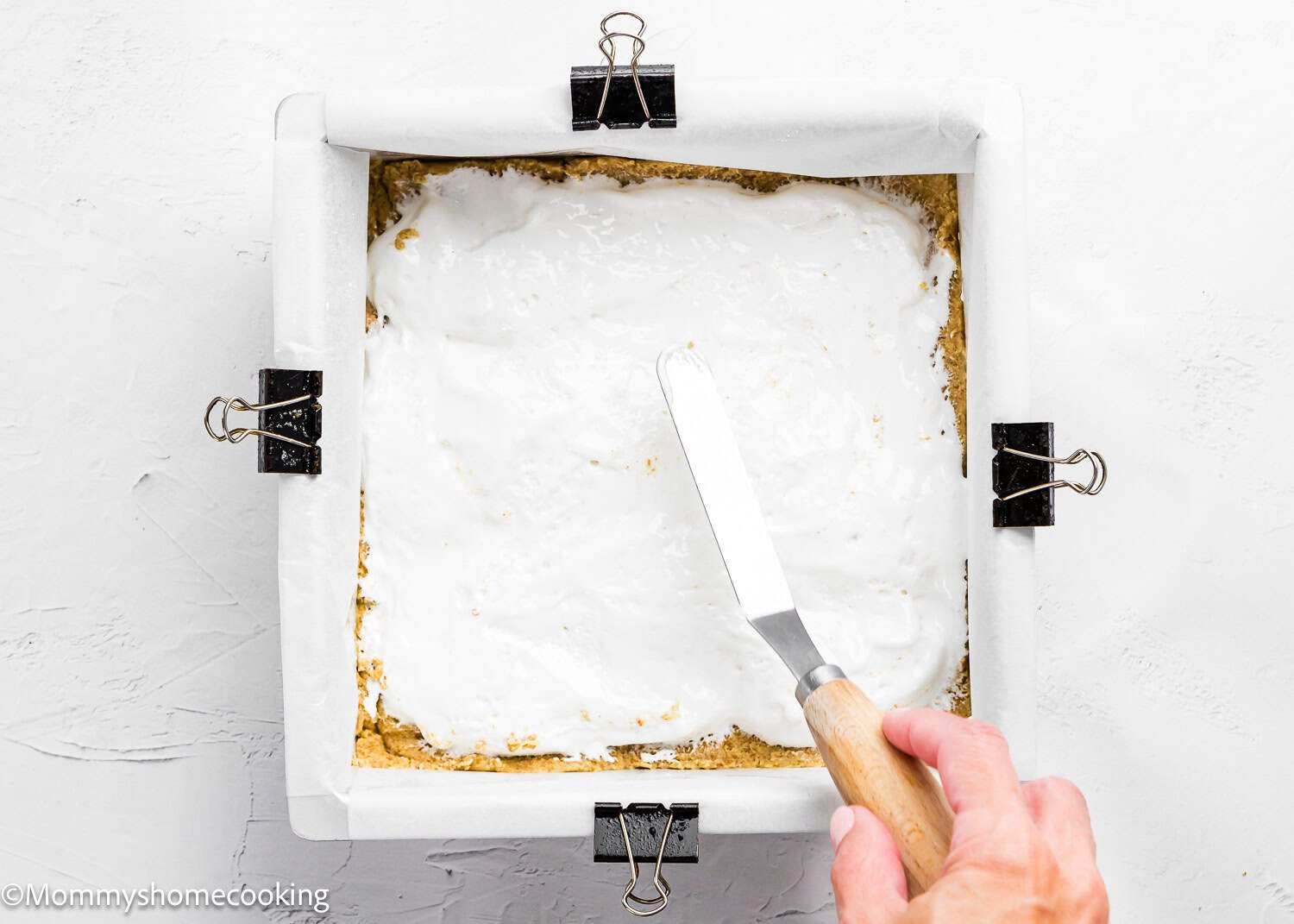 A hand spreads a white creamy layer over a crust in a parchment-lined square baking pan secured with binder clips, preparing Eggless S’mores Cookie Bars.