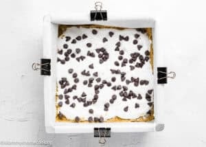 A square baking pan lined with parchment paper and clips, filled with Eggless S’mores Cookie Bars topped with marshmallow and scattered chocolate chips.