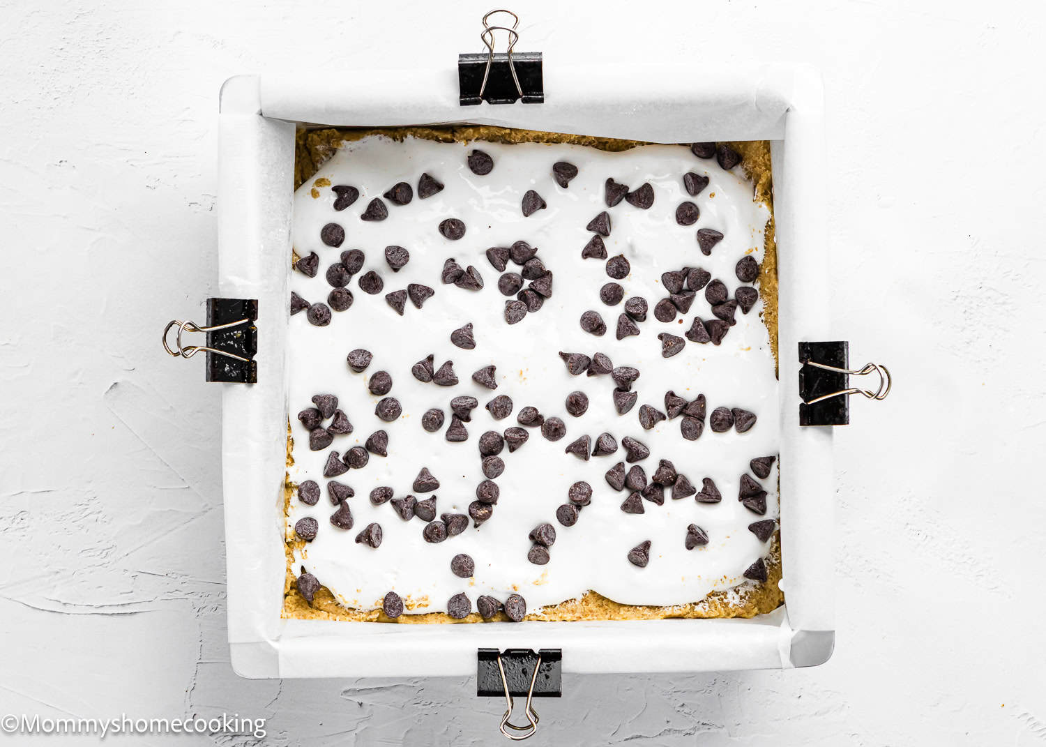 A square baking pan lined with parchment paper and clips, filled with Eggless S’mores Cookie Bars topped with marshmallow and scattered chocolate chips.