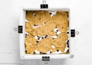 A square baking pan lined with parchment paper holds partially baked Eggless S’mores Cookie Bars, featuring cookie dough, marshmallow, and chocolate chips, secured with binder clips on each side.