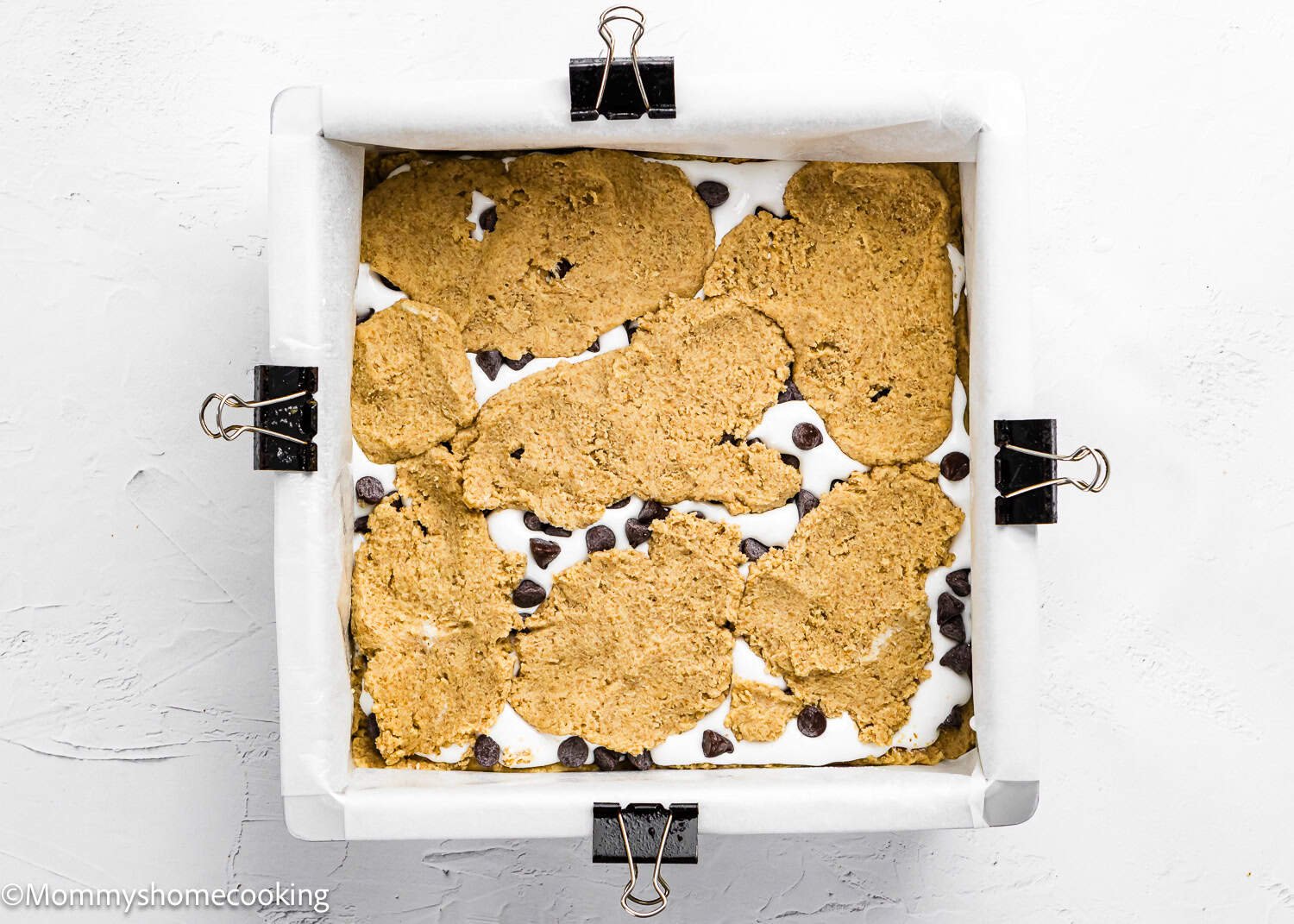 A square baking pan lined with parchment paper holds partially baked Eggless S’mores Cookie Bars, featuring cookie dough, marshmallow, and chocolate chips, secured with binder clips on each side.