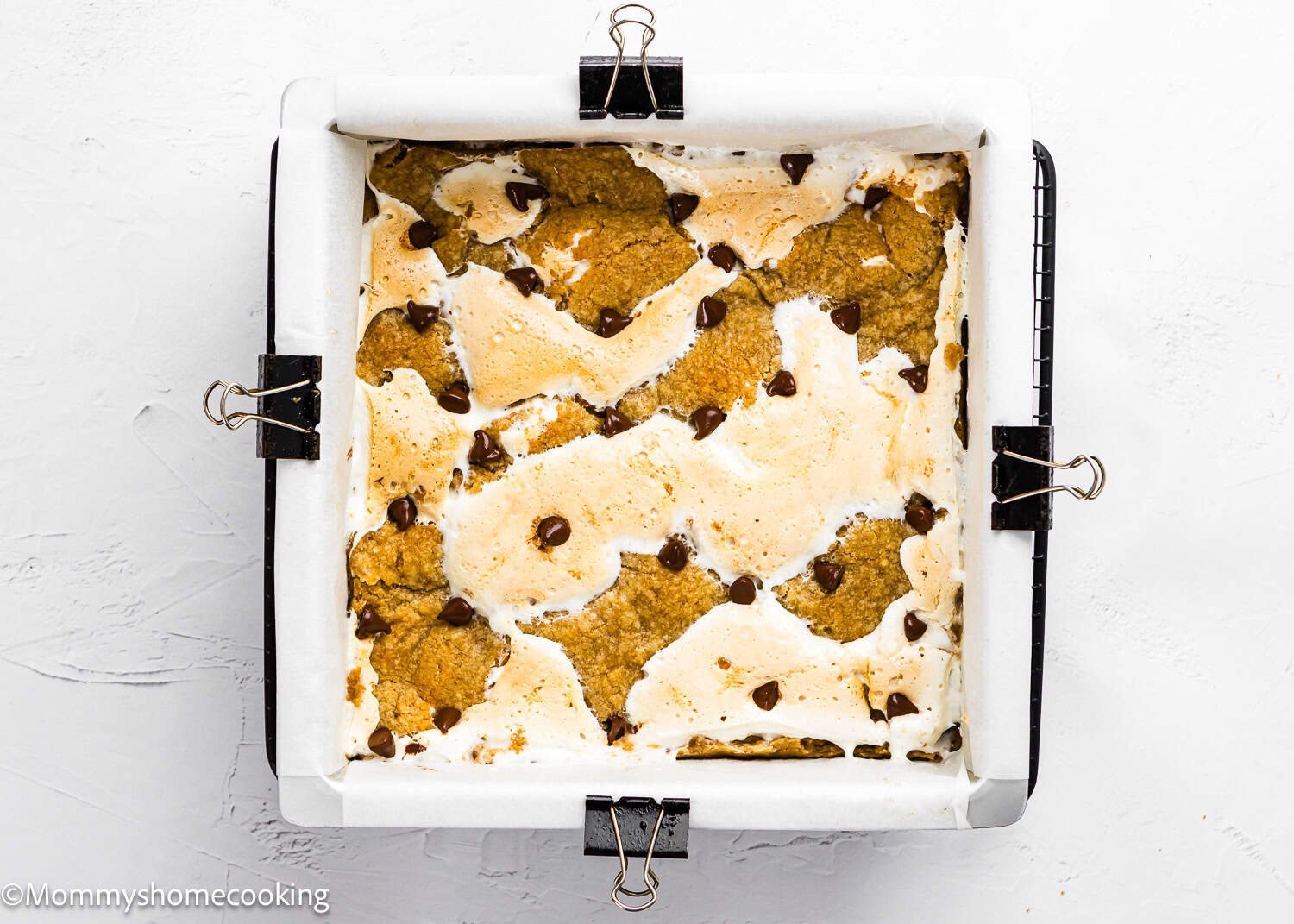 A square baking pan lined with parchment paper holds partially baked Eggless S’mores Cookie Bars topped with melted marshmallow and chocolate chips.