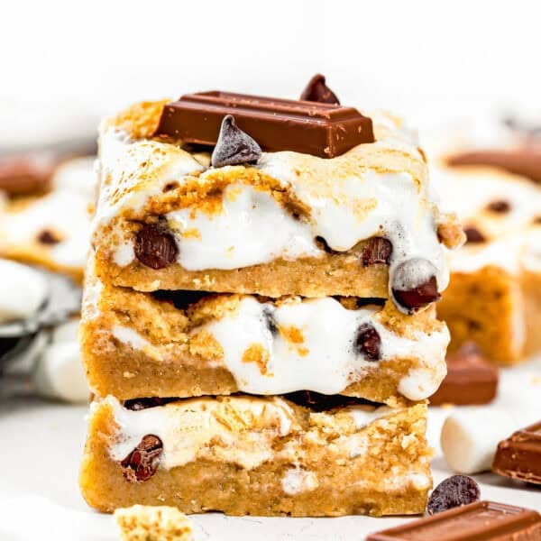 Three stacked Eggless S’mores Cookie Bars with visible chocolate chips, marshmallow, and chocolate pieces on top, set against a light background.