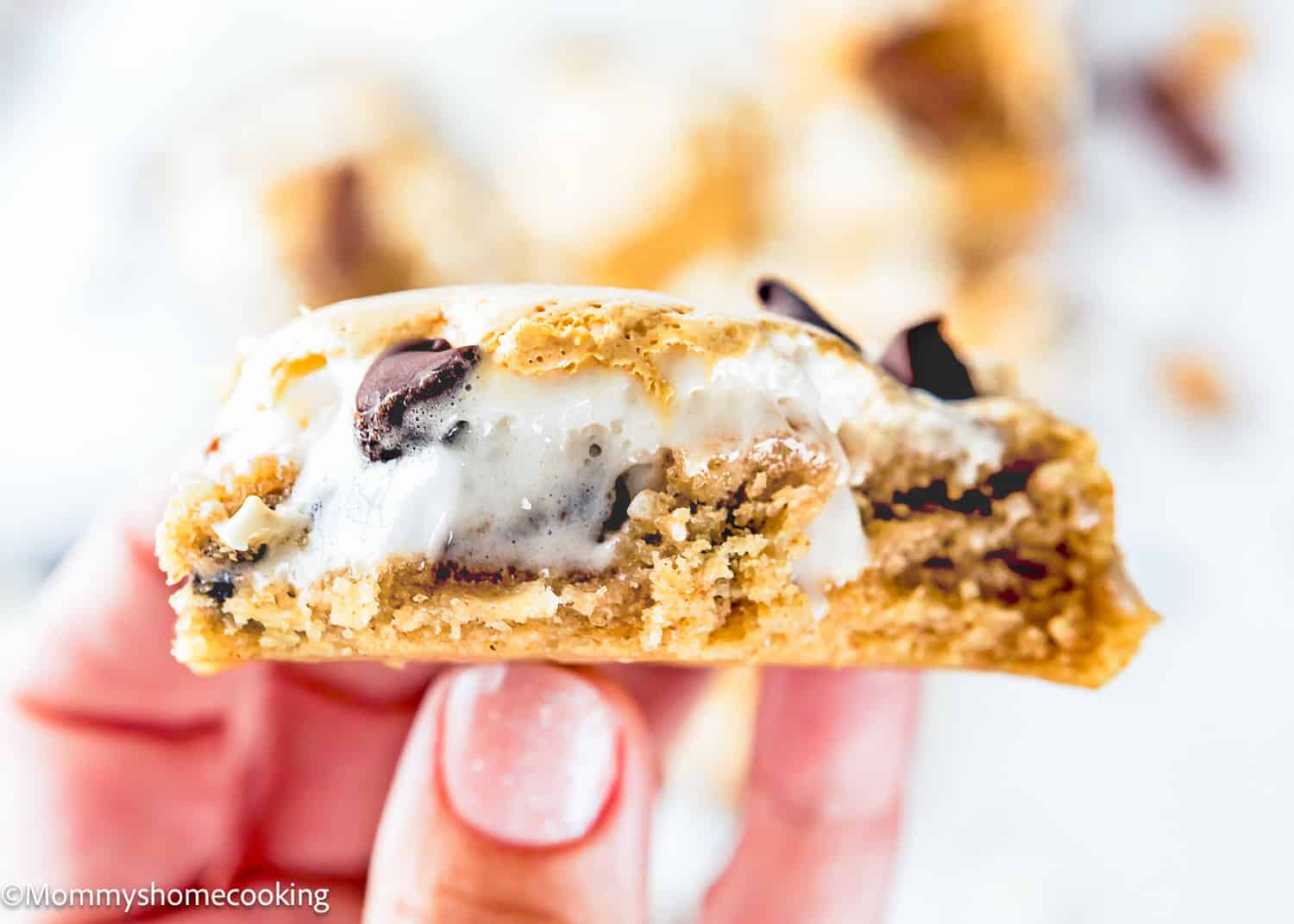 A hand holding a half-eaten Eggless S’mores Cookie Bar with visible chocolate chips and gooey marshmallow filling.