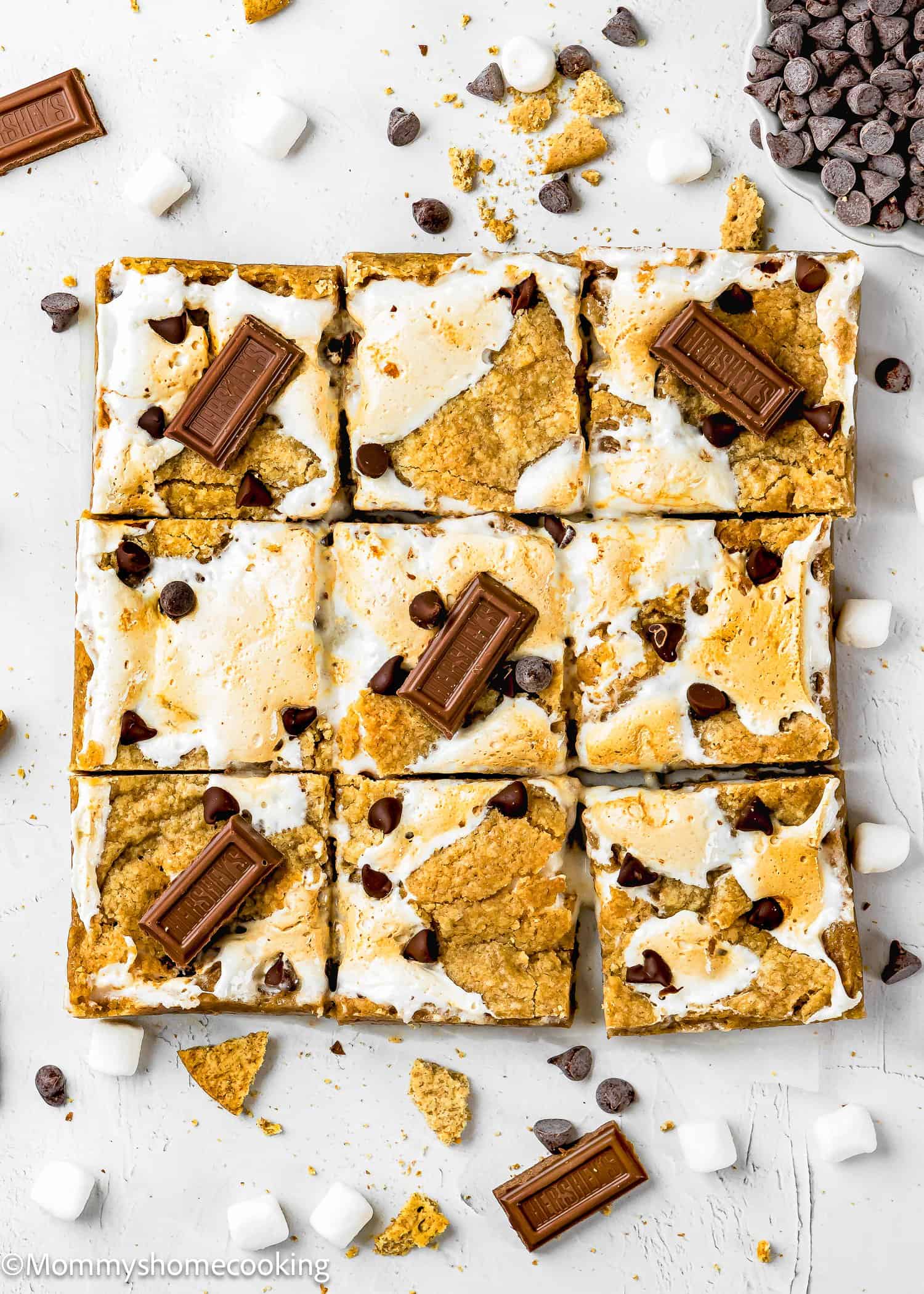 Nine Eggless S’mores Cookie Bars are arranged in a grid, topped with chocolate pieces, chocolate chips, and marshmallows on a white background with scattered crumbs and ingredients.