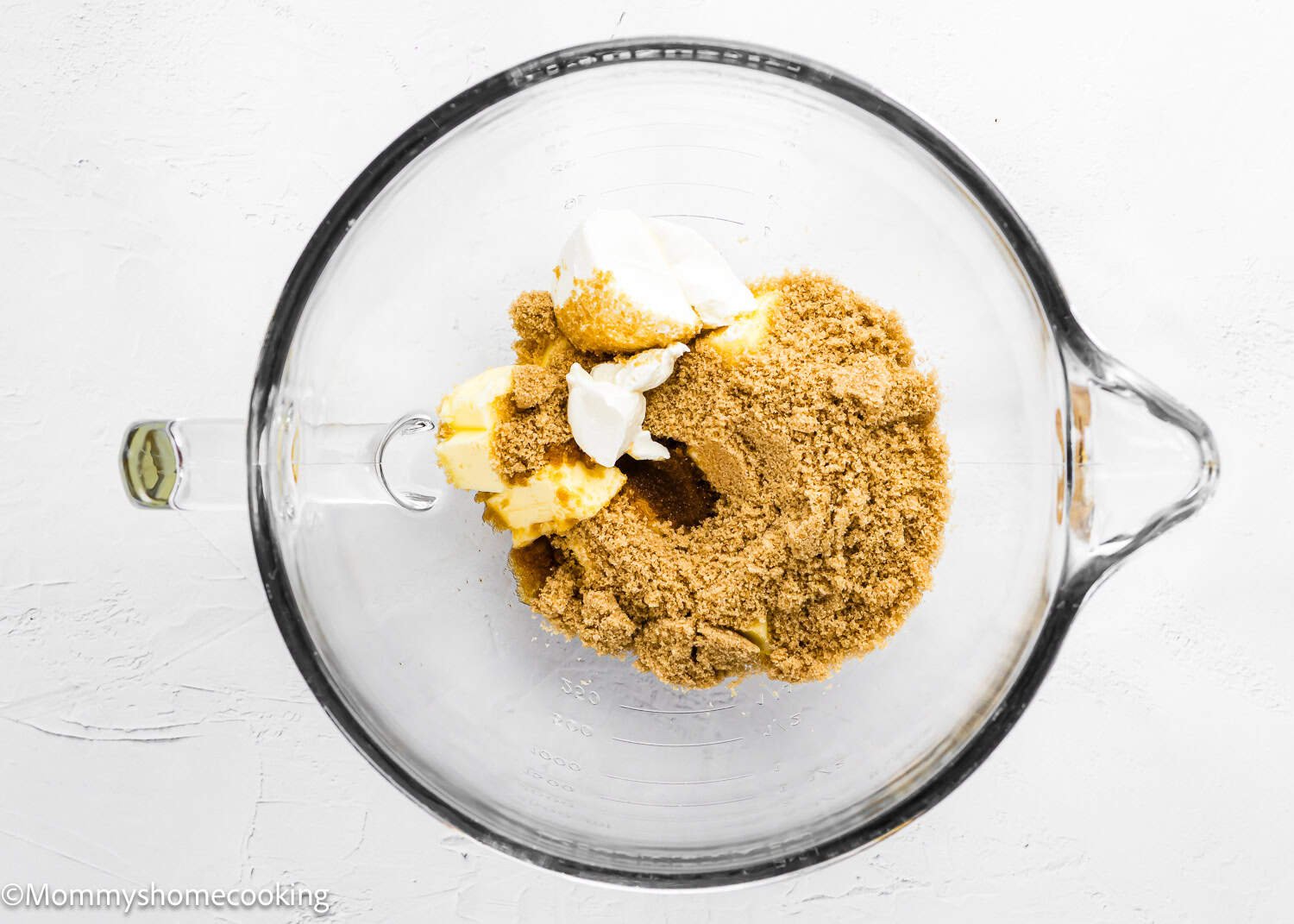A glass mixing bowl containing brown sugar, butter, a dollop of sour cream, and ingredients for Eggless S’mores Cookie Bars sits on a white surface.