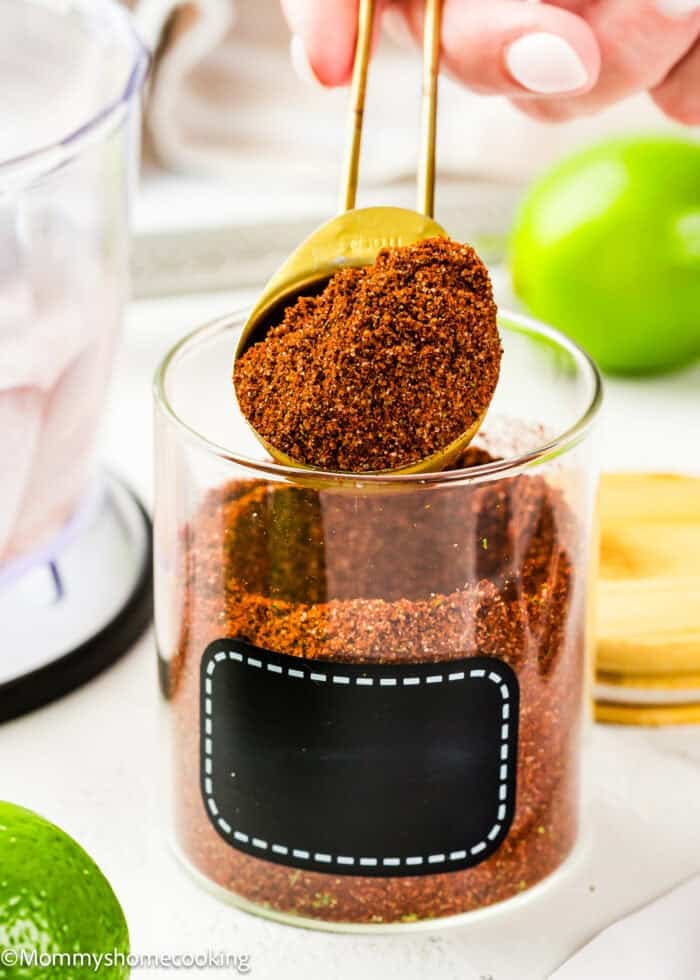 A hand holds a gold measuring spoon scooping Homemade Chili Lime Seasoning (No Additives & Allergy-Friendly) from a clear glass jar with a blank black label; a lime and wooden lid sit nearby.