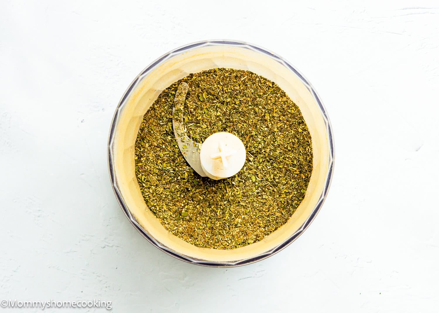 The food processor bowl contains finely ground green herbs on a white surface for homemade Greek seasoning blends (no filler, allergy-friendly).