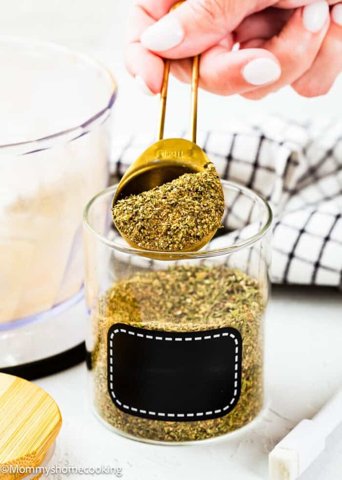 A hand holds a gold tablespoon of Homemade Greek Seasoning Blend (No Fillers,Allergy-Friendly) over a glass jar filled with the same seasoning.