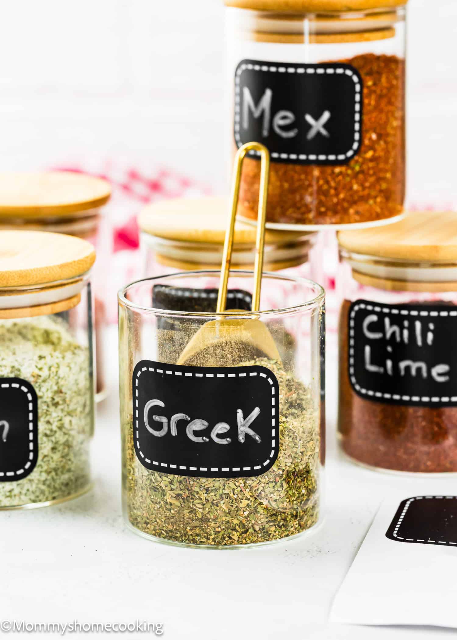 Glass spice jars with wooden lids and black labels display blends like "Homemade Greek Seasoning Blend (No Fillers, Allergy-Friendly)," "Chili Lime," and "Mex." A gold spoon rests in the jar labeled "Greek.